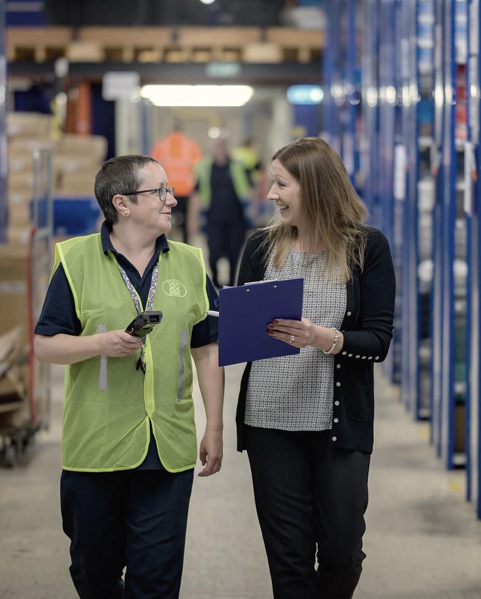 Two colleagues walking a warehouse aisle, one holding a clipboard, talking through the work.